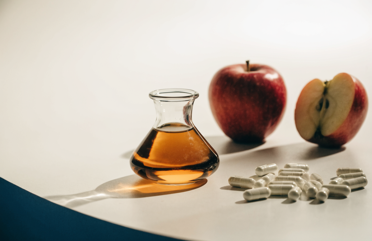 A glass carafe of amber apple cider vinegar beside white capsules and fresh apples