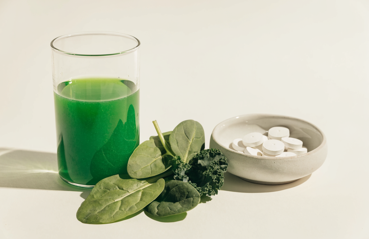 A glass of green drink beside fresh spinach and kale, with a dish of tablets alongside