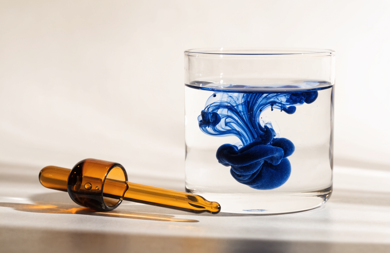 A drop of deep blue liquid dispersing through a clear glass of water beside a dropper
