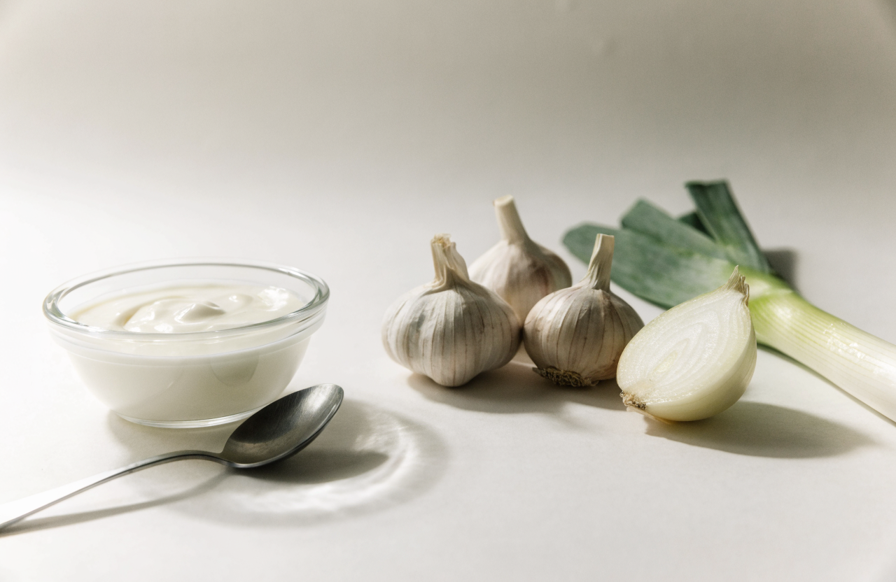 A bowl of plain yoghurt beside garlic, onion and leek on a pale surface